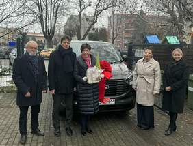 Pokaż zdjęcie: Grupa pięciu osób stoi przed samochodem z czerwona kokardą. W tle widoczne są drzewa bez liści oraz budynki. Osoby ubrane są w eleganckie, zimowe płaszcze.