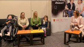 Pokaż zdjęcie: Na kanapach siedzą kobiety i dzieci. Rozmawiają ze sobą. Jedna z dziewczynek siedzi na elektrycznym wózku inwalidzkim. 