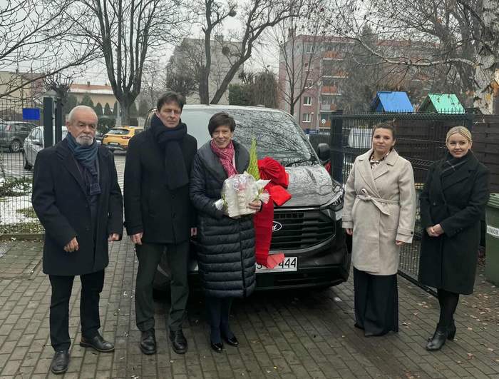 Pokaż zdjęcie: Grupa pięciu osób stoi przed samochodem z czerwona kokardą. W tle widoczne są drzewa bez liści oraz budynki. Osoby ubrane są w eleganckie, zimowe płaszcze.