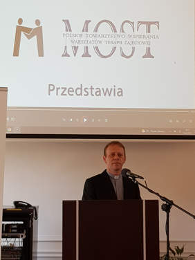 Ksiądz za mównicą, mikrofonem, nad nim napis MOST