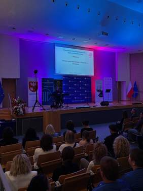Pokaż zdjęcie: Wypełniona publicznością aula. Uczestnicy siedzą na widowni i obserwują scenę, na której znajduje się osoba przy instrumencie. W tle widoczny jest ekran z prezentacją.