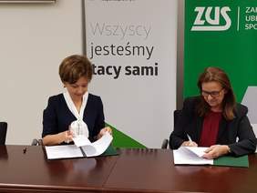 Pokaż zdjęcie: Dwie kobiety siedzą przy stole. W rękach trzymaja długopisy, którymi podpisuja dokumenty leżące na stole