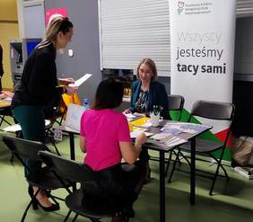 Pokaż zdjęcie: Stoisko informacyjne PFRON. Przy stoliku rozmawiają trzy osoby, na stoliku wyeksponowane są materiały informacyjne.