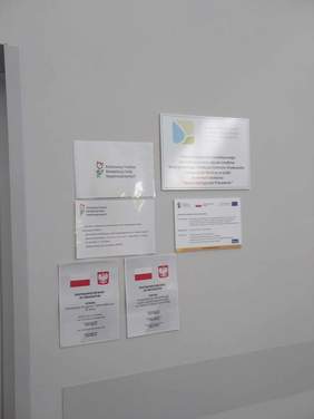 Pokaż zdjęcie: Na białej ścianie wiszą tablice z informacjami o dofinansowaniach Państwowego Funduszu Rehabilitacji Osób Niepełnosprawnych. 