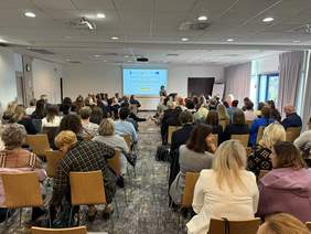 Pokaż zdjęcie: ala konferencyjna wypełniona uczestnikami siedzącymi w rzędach, skierowanymi w stronę mównicy i ekranu prezentacyjnego z wyświetlonym slajdem. Pomieszczenie jest jasne, z dużymi oknami po prawej stronie i zasłonami.