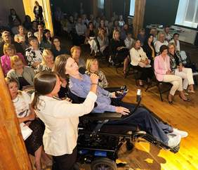 Pokaż zdjęcie: Grupa kilkudziesięciu osób siedzi na sali i przysłuchuje się wystąpieniu kobiety poruszającej się na wózku. Druga kobieta stoi przy prelegentce, trzymając jej mikrofon, z którego korzysta.