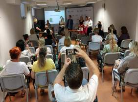 Pokaż zdjęcie: spotkanie przedstawicieli powiatów i pracowników PFRON w tle duży telebim na którym wyświetlane są slajdy obok stoi pracownik Funduszu i przeproszadza rozmowę. Na dużej sali siedzą osoby które pokazane są z tyłu.