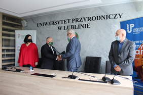 Pokaż zdjęcie: W sali konferncyjnej dwaj mężczyźni stojąc podaja sobie dłonie.Oprócz nich stoi jeden mężczyzna i jedna kobieta.W tle loga Uniwersytetu i PFRON