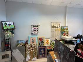 Pokaż zdjęcie: Fragment pomieszczenia, na tle szarej ściany w trzech rzędach ustawione są kolorowe obrazy.