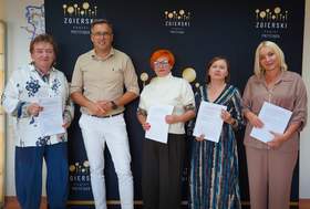Do zdjęcia pozuje mężczyzna i cztery kobiety. Panie prezentują podpisane umowy. W tle widać baner powiatu zgierskiego. 