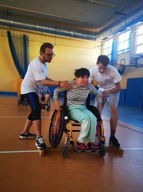 Pokaż zdjęcie: na zdjęciu hala sportowa, osoba poruszająca się na wózku uczy się podjazdu z przeszkodą obok niej stoją dwaj instruktorzy którzy pomagają przy manewrach 