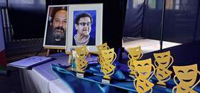 Pokaż zdjęcie: Na stole stoją statuetki - nagrody w Ogólnopolskim Festiwalu Twórczości Teatralno-Muzycznej Osób Niepełnosprawnych Intelektualnie Albertiana 2025. W tle stoją także portrety dwóch osób: Bożenny Piotrowicz oraz księdza Tadeusza Isakowicza-Zaleskiego.