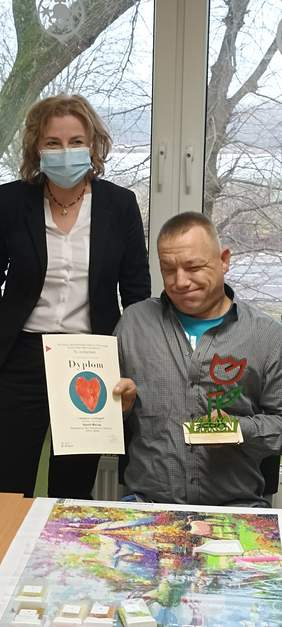 Pokaż zdjęcie: .Dwie postacie, kobieta w maseczce i mężczyzna trzymający w prawej dłoni dyplom na którym widzimy czerwone serce na niebieskim kółku, w lewej dłoni statuetkę przedstawiającą czerwono-zielonego kwiatka .