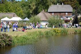 Pokaż zdjęcie: Na pierwszym planie staw, za nim budynku skansenu, na trawie siedzą uczestnicy pikniku lub stoją i rozmawiają w grupach, słoneczna pogoda 