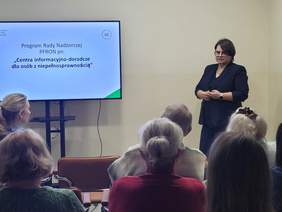 Pokaż zdjęcie: Prezentacja dotycząca CIDON. Na środku sali ekran z prezentacją. Obok ekranu stoi kobieta, która przemawia do osób niedzących na krzesłach. 