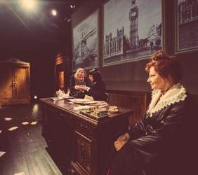 Pokaż zdjęcie: Fragment sceny na której grane jest przedstawienie. Jedna aktorka siedzi z prawej strony biurka, druga siedzi za biurkiem nad nią nachyla się kobieta i pokazując dłonie coś jej tłumaczy.