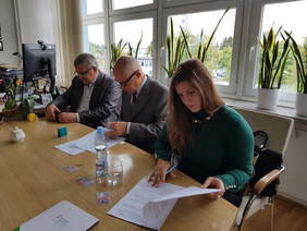 Pokaż zdjęcie: Trzy osoby, dwóch mężczyzn i kobieta siedzą przy jasnym dużym stole nad dokumentami, za nimi ściana okien