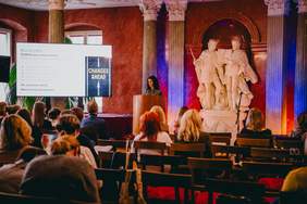 Pokaż zdjęcie: Sala konferencyjna z publicznością siedzącą na krzesłach i zwróconą w stronę sceny.  Na środku widać mównicę, za którą stoi osoba prowadząca prezentację.  Po lewej stronie sceny ustawiony jest ekran projekcyjny i wyświetlony jest na nim slajd. Za sceną znajdują się wysokie kolumny oraz duża jasna rzeźba przedstawiająca dwie stojące postacie ludzkie na postumencie.  Ściany mają ciepły, czerwony kolor, a oświetlenie w odcieniach pomarańczowo niebieskich podkreśla kolumny i rzeźbę.  
