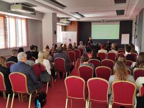 Widok na salę z tyłu - na krzesłąch siedzą słuchacze, a na przodzie prowadzący