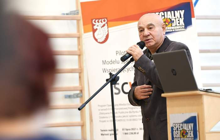 Pokaż zdjęcie: Przy mikrofonie stoi starszy mężczyzna w garniturze. W tle rollupy z logo Specjalnego Ośrodka Szkolno Wychowawczego.