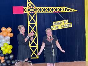 Pokaż zdjęcie: Na środku stoją dwie kobiety. Jedna przemawia do mikrofonu, a druga bije brawo. W tle jest granatowa ściana z rysunkami dźwigu budowlanego. 