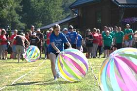 Pokaż zdjęcie: Kobieta w jednokolorowej koszulce i spodenkach biegnie za dużą półprzezroczystą piłką w kolorowe pasy za nią kibice i mężczyzna biegnący z piłką w przeciwną stronę.