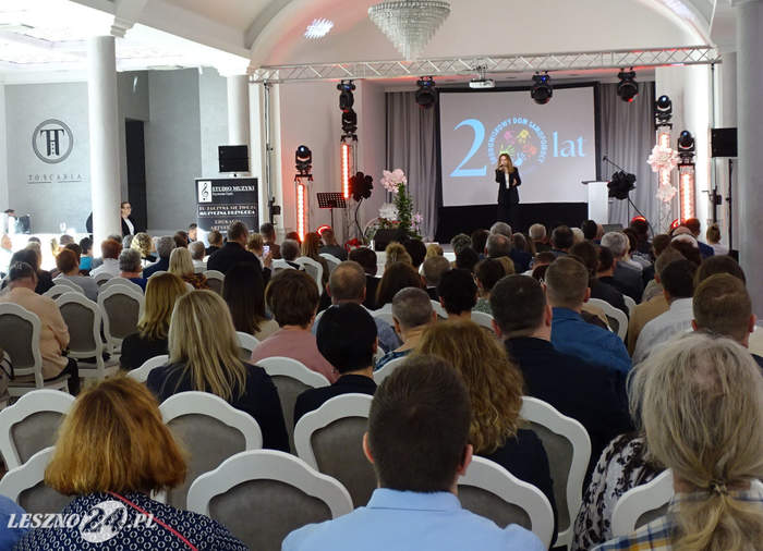 Pokaż zdjęcie: W dużej sali znajduje się grupa kilkudziesięciu osób, które siedzą w kilku rzędach. Z przodu stoi na scenie kobieta z mikrofonem, a za nią duży ekran z napisem informującym o jubileuszu 20-lecia Środowiskowego Domu Samopomocy. 