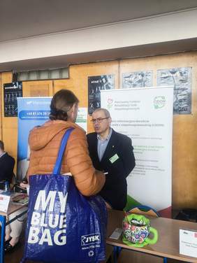 Pokaż zdjęcie: Osoba odwiedzająca rozmawia z przedstawicielem Oddziału Lubelskiego PFRON przy stoisku informacyjnym podczas Targów Pracy. W tle widoczny roll-up PFRON i plakat Centrum Informacyjno-Doradczego.