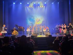 Pokaż zdjęcie: Na scenie występują artyści ze Studia Integracji. Niektórzy stoją, a inni siedzą na krzesłach i wózkach inwalidzkich. Na widowni siedzi publiczność. 