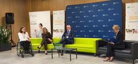 Pokaż zdjęcie: Trwa panel dyskusyjny konferencji „Dostępność dziś i jutro”. Rozmawiają trzy kobiety i jeden mężczyzna. Jedna z pań siedzi na ręcznym wózku inwalidzkim. W tle widać banery różnych instytucji. 