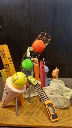 Pokaż zdjęcie: Barwna i intensywna kolorystyka. Na środku żółto brunatna, kwadratowa podstawka. Na niej po lewej stronie dwie piłki: pomarańczowoczerwona i jaskrawa zieleń. W prawym rogu sześć niebiesko jasnych, modrych, czerwonych małych samochodzików. Po dr