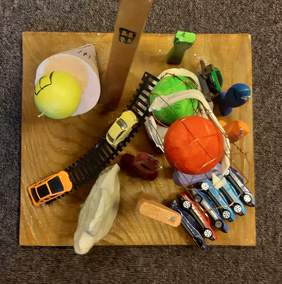 Pokaż zdjęcie: Barwna i intensywna kolorystyka. Na środku żółto brunatna, kwadratowa podstawka. Na niej po lewej stronie dwie piłki: pomarańczowoczerwona i jaskrawa zieleń. W prawym rogu sześć niebiesko jasnych, modrych, czerwonych małych samochodzików. Po dr