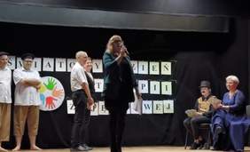 Pokaż zdjęcie: Uczestnicy WTZ prezentują na scenie umiejętności aktorskie. Dwóch mężczyzn po lewej stronie w białych koszulach, spodniach 3/4 i na boso, a po prawej stronie na krzesłach siedzi kobieta i mężczyzna. W tle czarny napis na białym tle "Światowy Dzień Warsztatów Terapii Zajęciowej" oraz grafika składające się z kolorowych dłoni ułożonych w okrag.