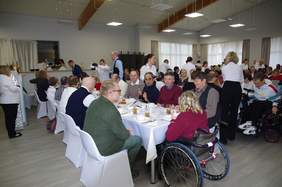 Pokaż zdjęcie: Duża sala podczas wspólnego posiłku, w której przy długich stołach przykrytych białymi obrusami siedzi kilkadziesiąt osób. Wśród gości znajdują się osoby poruszające się na wózkach inwalidzkich, w pełni zintegrowane z pozostałymi uczestnikami. W tle widać personel obsługujący wydarzenie w białych koszulach oraz bufet. Pomieszczenie jest przestronne, z nowoczesnym oświetleniem sufitowym i dużymi oknami z jasnymi zasłonami.