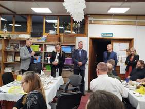 Pokaż zdjęcie: Grupa osób uczestniczących w spotkaniu wigilijnym organizowanym w Warsztatach Terapii Zajęciowej w Ogrodniczkach. Uczestnicy siedzą przy stole ustawionym w kształcie litery „U”, na którym znajdują się drobne poczęstunki i filiżanki. Kilka osób stoi w tle, w tym jedna z osób  trzyma torbę z podarunkami i przemawiające do zgromadzonych. Wśród przemawiających jest mężczyzna, który wręcza symboliczny upominek. W tle widać regały z różnorodnym wyposażeniem i materiałami warsztatowymi oraz dekoracje.