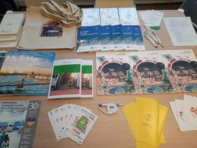 Pokaż zdjęcie: Na stoliku znajdują się różne materiały promocyjne i reklamowe PFRON. Są to różnego rodzaju: poradniki, opracowania, broszury, ulotki, długopisy i torby. 