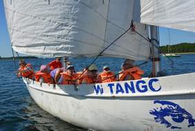 Pokaż zdjęcie: Dziesięć osób ubranych w pomarańczowe kapoki płynie białym jachtem po jeziorze.