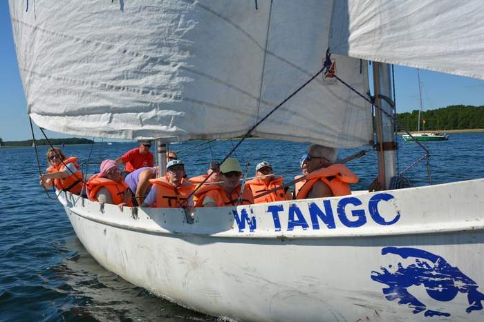 Pokaż zdjęcie: Dziesięć osób ubranych w pomarańczowe kapoki płynie białym jachtem po jeziorze.