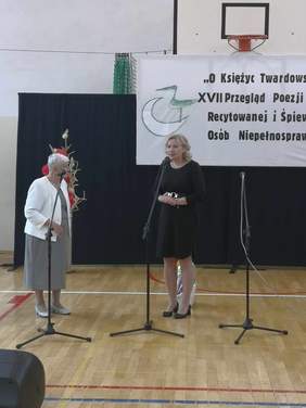 Pokaż zdjęcie: Na scenie przy mikrofonie stoi kobieta, blondynka w czarneh sukience.