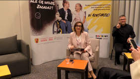 Pokaż zdjęcie: Na fotelu siedzi elegancko ubrana kobieta. Po prawej stronie znajduje się tłumacz języka migowego. W tle widać plakat reklamowy kampanii społecznej „W kontakcie”. 