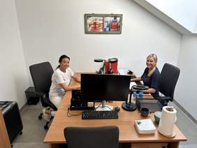 Pokaż zdjęcie: Przy biurkach naprzeciwko siedzą dwie uśmiechnięte kobiety. Na biurkach stoją monitory komputerowe.