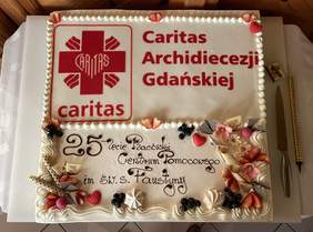Pokaż zdjęcie: Kremowy tort w prostokątnym kształcie zdobiony lukrowymi kwiatami po bokach. W górnej połowie logo Caritas w dolnej napis 25 lecie Centrum Pomocowe im. św. siostry Faustyny.