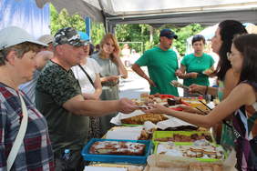 Pokaż zdjęcie: Pod namiotem stoisko gastronomiczne, dwie kobiety nakładają gościom porcje ciasta