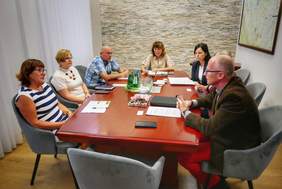 Pokaż zdjęcie: Siedem osób siedzi wokół dużego, prostokątnego stołu konferencyjnego w gabinecie. Wszyscy są skupieni na rozmowie. Na stole leżą dokumenty. Pomieszczenie ma ściany z naturalnego kamienia i jasne zasłony. Na ścianie w tle wisi oprawiona mapa lub plakat.