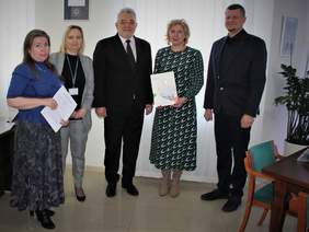 Pokaż zdjęcie: Trzy kobiety i  dwaj mężczyźni stoją w półokręgu. Dwie z kobiet trzymają w ręku kartki.