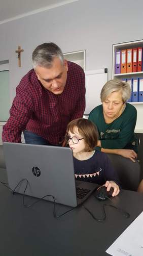 Pokaż zdjęcie: Przy laptopie siedzi dziewczynka, nad nią stoją dwie osoby, mężczyzna i kobieta, tłumaczą jej obsługę komputera