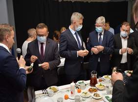 Pokaż zdjęcie: Grupa osób ubrana odświętnie w garnitury w maseczkach dzieli się opłatkiem przy stole  z potrawami wigilijnymi 