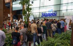 Pokaż zdjęcie: Do rejestracji w budynku stoi długa kolejka osób. Nad punktem rejestracyjnym napis solidarność, dookoła dużo zieleni.