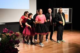 Pokaż zdjęcie: Na scenie tańczą 3 pary, mężczyźni ubrani na czarno, kobiety na czarno czerwono. 