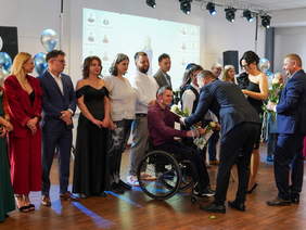 Pokaż zdjęcie: Grupa osób ubrana elegancko stoi na scenie. Na pierwszym planie widać mężczyznę na wózku, który otrzymuje kwiaty i podziękowania za wspólny wkład w historię Zakładu Aktywności Zawodowej. Pozostali uczestnicy, zarówno kobiety jak i mężczyźni, stoją w rzędach, również trzymając kwiaty. W tle widoczne są dekoracje w postaci balonów oraz oświetlenie sceniczne. 